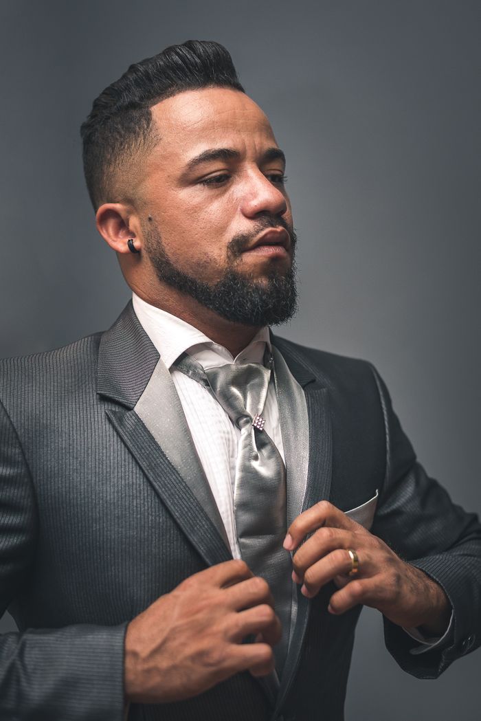 Formal portrait of a bearded man adjusting his tie in a gray suit against a plain backdrop.