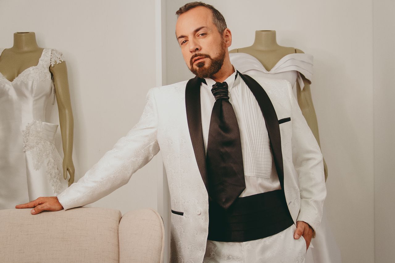 A man in a stylish white tuxedo stands confidently in a studio, surrounded by mannequins.
